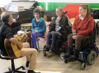 Music therapist Allan Slade on guitar and (left to right) Kaitlin, Corrine and Sierra. Corrine and Sierra play instruments using the iPods attached to their arms, while Katlin presses a grey accessibility switch.